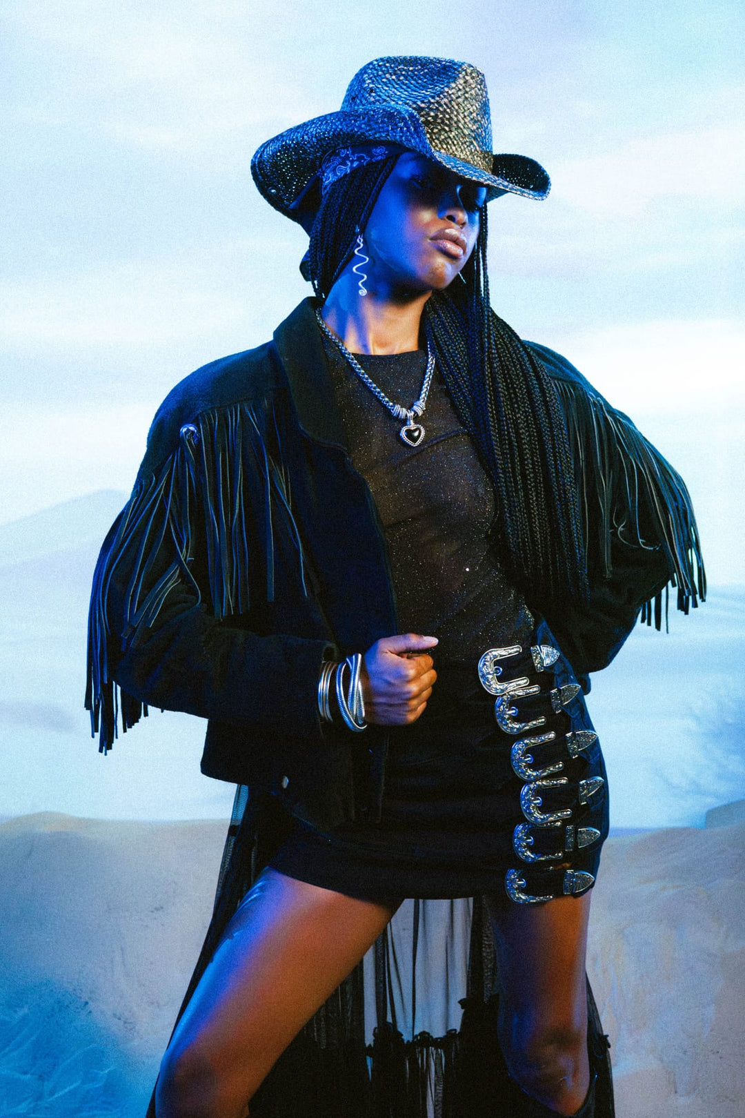 Model in black suede fringe jacket and cowboy hat with silver buckle belt against a blue-hour studio backdrop