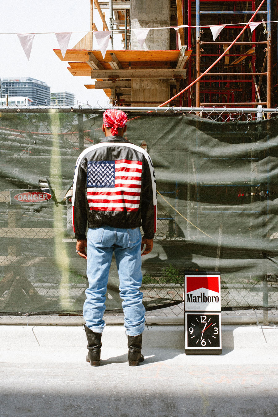 From behind: a leather USA flag jacket, light jeans and cowboy boots by a construction fence